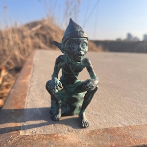 Puede incluir: Una escultura de bronce de un elfo sentado, con orejas puntiagudas y un gorro puntiagudo. El elfo está en cuclillas con las manos sobre las rodillas. La escultura tiene un aspecto envejecido y está sobre un fondo borroso al aire libre.
