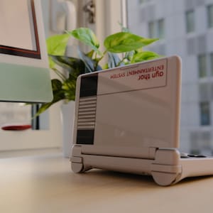 May include: A white handheld gaming console with a retro design, featuring a screen and a keyboard. The console has the text "Ayn Thor Entertainment System" on the top. It is placed on a light-colored surface near a window with a plant and a computer.