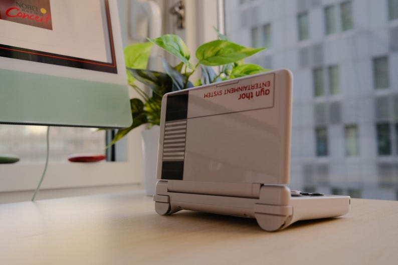 May include: A white handheld gaming console with a retro design, featuring a screen and a keyboard. The console has the text "Ayn Thor Entertainment System" on the top. It is placed on a light-colored surface near a window with a plant and a computer.