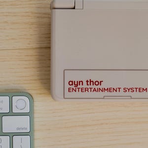 May include: A light beige entertainment system with the text "ayn thor ENTERTAINMENT SYSTEM" in red. A light green and white keyboard is in the lower left corner. The objects are on a light wood surface.