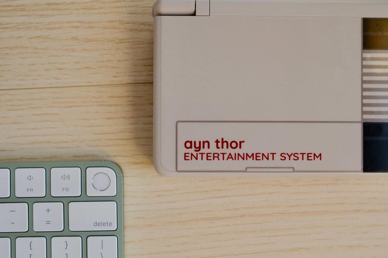May include: A light beige entertainment system with the text "ayn thor ENTERTAINMENT SYSTEM" in red. A light green and white keyboard is in the lower left corner. The objects are on a light wood surface.