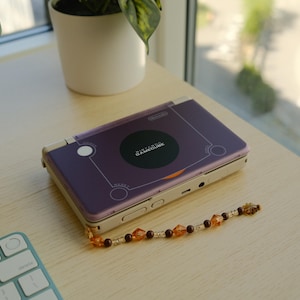 May include: A purple Nintendo GameCube handheld console with a decorative bead chain. The console has a black circular design with the words "GameCube" printed on it. The console is on a light-colored wooden surface.