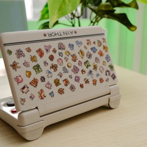 May include: A white handheld gaming console with a colorful array of cartoon character stickers. The console is open, displaying the screen with the stickers. The text "AYN THOR" is printed on the top of the console.