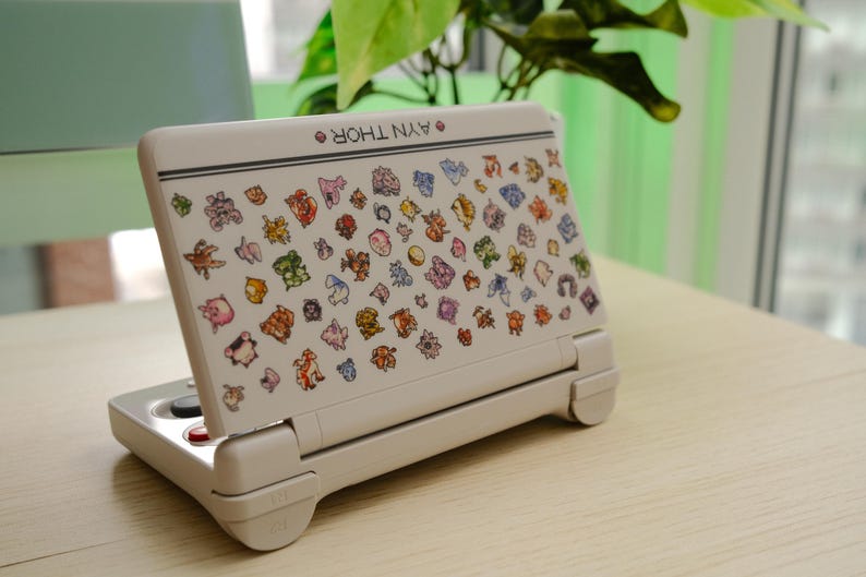 May include: A white handheld gaming console with a colorful array of cartoon character stickers. The console is open, displaying the screen with the stickers. The text "AYN THOR" is printed on the top of the console.