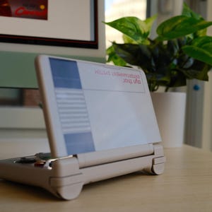 May include: A handheld gaming device with a white and blue screen, and a beige base. The device is open, displaying text that reads "ENTERTAINMENT SYSTEM" and "ayn thor". A potted plant is in the background.