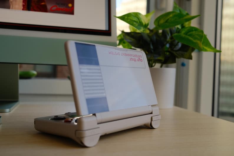 May include: A handheld gaming device with a white and blue screen, and a beige base. The device is open, displaying text that reads "ENTERTAINMENT SYSTEM" and "ayn thor". A potted plant is in the background.
