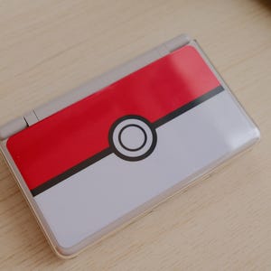 May include: A Nintendo DS Lite handheld gaming console with a custom skin featuring a red and white Pokeball design. The top half is red, the bottom half is white, and a black circle with a white center is in the middle.