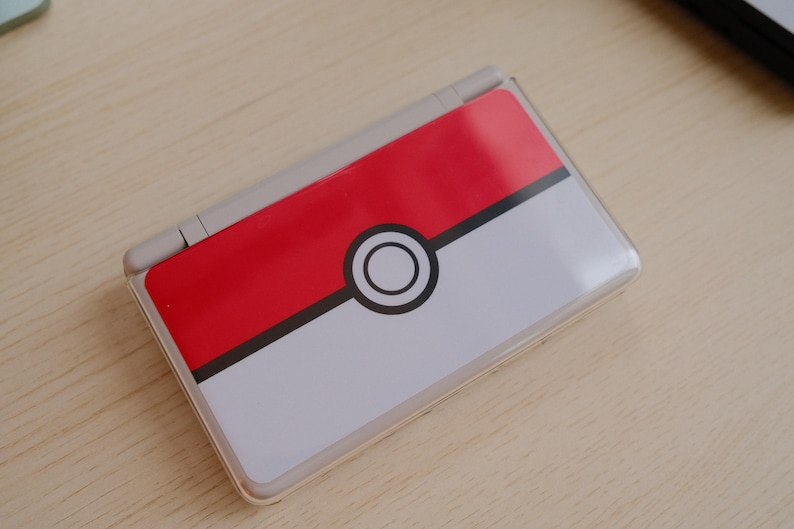 May include: A Nintendo DS Lite handheld gaming console with a custom skin featuring a red and white Pokeball design. The top half is red, the bottom half is white, and a black circle with a white center is in the middle.