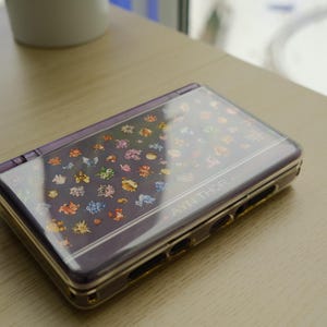 May include: A purple handheld gaming console with a clear cover featuring colorful cartoon characters. The console is closed and resting on a light-colored wooden surface. The text "AYN THOR" is visible on the device.