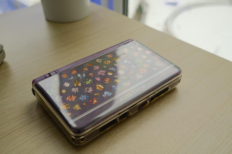 May include: A purple handheld gaming console with a clear cover featuring colorful cartoon characters. The console is closed and resting on a light-colored wooden surface. The text "AYN THOR" is visible on the device.