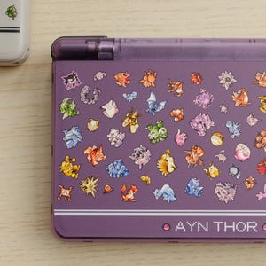 May include: Purple handheld gaming console with a clear top and a design of colorful cartoon characters. The text "AYN THOR" is printed on the bottom of the console.