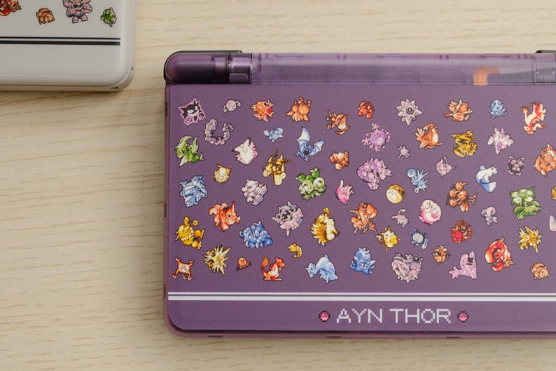 May include: Purple handheld gaming console with a clear top and a design of colorful cartoon characters. The text "AYN THOR" is printed on the bottom of the console.