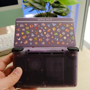 May include: A purple handheld gaming console with a decorated top featuring colorful character illustrations. The console is open, revealing the screen and buttons. The text "AYN THOR" is visible on the top. A second console is visible in the background.
