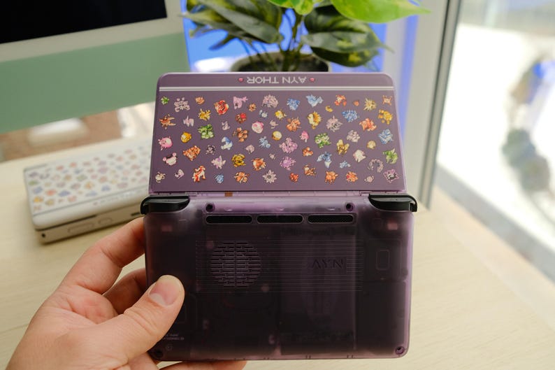 May include: A purple handheld gaming console with a decorated top featuring colorful character illustrations. The console is open, revealing the screen and buttons. The text "AYN THOR" is visible on the top. A second console is visible in the background.