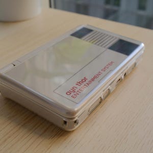 May include: A silver handheld gaming console with the text "ayn thor ENTERTAINMENT SYSTEM" in red. The console has a rectangular shape with rounded corners and a hinged design. The top of the console features a graphic design with black, white, and yellow stripes.