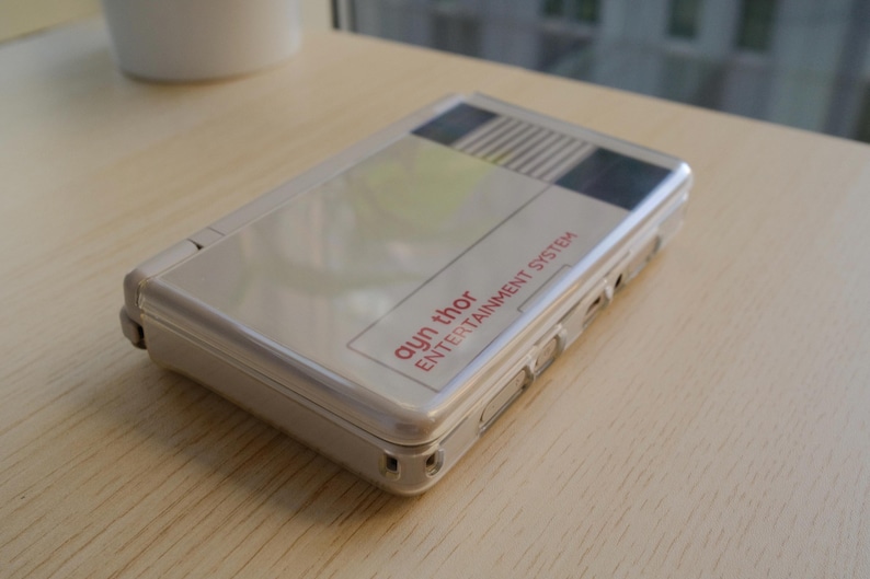 May include: A silver handheld gaming console with the text "ayn thor ENTERTAINMENT SYSTEM" in red. The console has a rectangular shape with rounded corners and a hinged design. The top of the console features a graphic design with black, white, and yellow stripes.