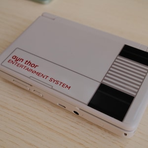 May include: A handheld gaming device in a light gray color, featuring the text "ayn thor ENTERTAINMENT SYSTEM" in red. The design includes black and gray striped accents, resembling a classic console.
