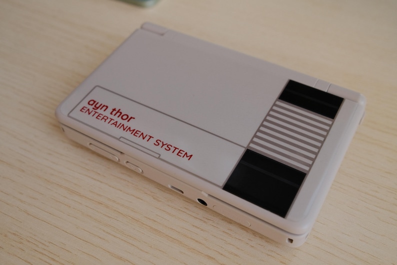 May include: A handheld gaming device in a light gray color, featuring the text "ayn thor ENTERTAINMENT SYSTEM" in red. The design includes black and gray striped accents, resembling a classic console.
