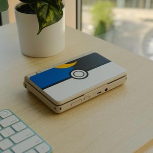 May include: A beige handheld gaming console with a custom skin featuring a split design: blue and yellow on one side, black and white with a circular graphic on the other. The console is resting on a light wood surface.