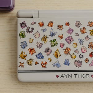 May include: A white handheld gaming console with a colorful pattern of various cartoon characters. The text "AYN THOR" is printed in pink at the bottom. A purple console is partially visible in the background.