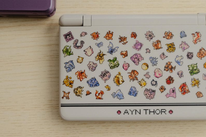 May include: A white handheld gaming console with a colorful pattern of various cartoon characters. The text "AYN THOR" is printed in pink at the bottom. A purple console is partially visible in the background.