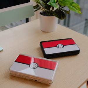 May include: Two handheld gaming consoles with a red, white, and black design, resembling a Pok&eacute; Ball, rest on a light-colored wooden surface. The consoles are open and closed, with a plant and computer monitor in the background.