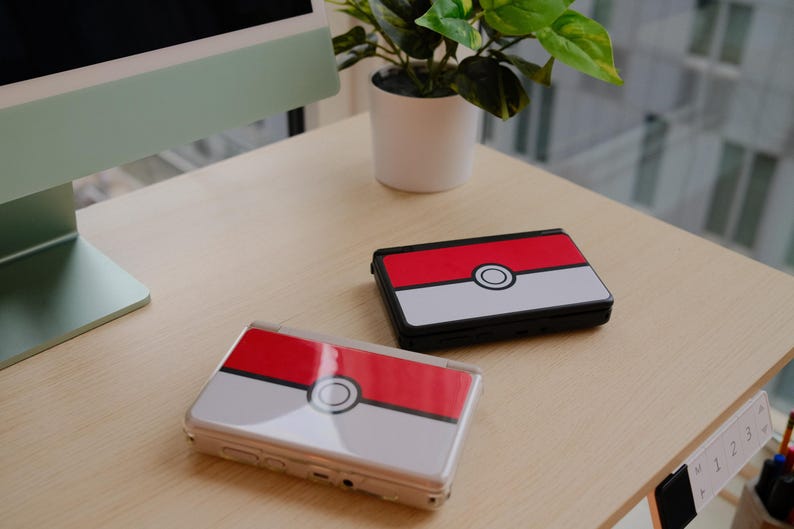 May include: Two handheld gaming consoles with a red, white, and black design, resembling a Pok&eacute; Ball, rest on a light-colored wooden surface. The consoles are open and closed, with a plant and computer monitor in the background.