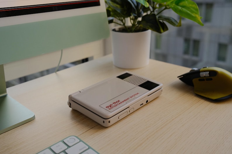Ayn Thor themed with a decal inspired by Nintendo Entertainment System console on desk, soft lighting, green computer mouse and plant in background.