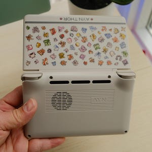 May include: A handheld gaming console in light gray with a screen featuring colorful cartoon characters. The console's back has a speaker and vents. The text "AYN THOR" is visible on the top.
