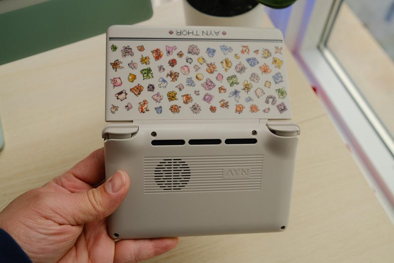 May include: A handheld gaming console in light gray with a screen featuring colorful cartoon characters. The console's back has a speaker and vents. The text "AYN THOR" is visible on the top.