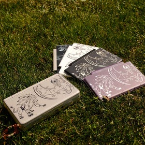 Puede incluir: Varias consolas de juegos portátiles en tonos plateado, negro, blanco y morado se muestran sobre hierba verde. Cada consola tiene un diseño decorativo de león. Las consolas están cerradas, lo que indica que no están en uso.