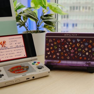 May include: A white Nintendo 3DS handheld game console with the "Pokémon Red Version" logo displayed on the screen. A purple case with a pattern of colorful characters is next to the console. The screen shows a Charmander character.