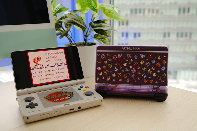 May include: A white Nintendo 3DS handheld game console with the "Pok&eacute;mon Red Version" logo displayed on the screen. A purple case with a pattern of colorful characters is next to the console. The screen shows a Charmander character.