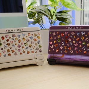 May include: Two handheld gaming consoles, one white and one purple, are displayed side-by-side. Both consoles feature colorful character graphics and the text "AYN THOR". The white console has a light gray base, while the purple one has a transparent purple base.