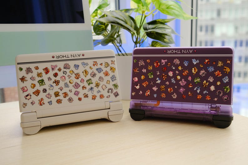 May include: Two handheld gaming consoles, one white and one purple, are displayed side-by-side. Both consoles feature colorful character graphics and the text "AYN THOR". The white console has a light gray base, while the purple one has a transparent purple base.