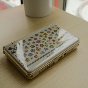 May include: A silver handheld gaming console with a clear cover featuring colorful cartoon character graphics. The console is open and resting on a light-colored wooden surface. The text "AYN THOR" is visible on the side.