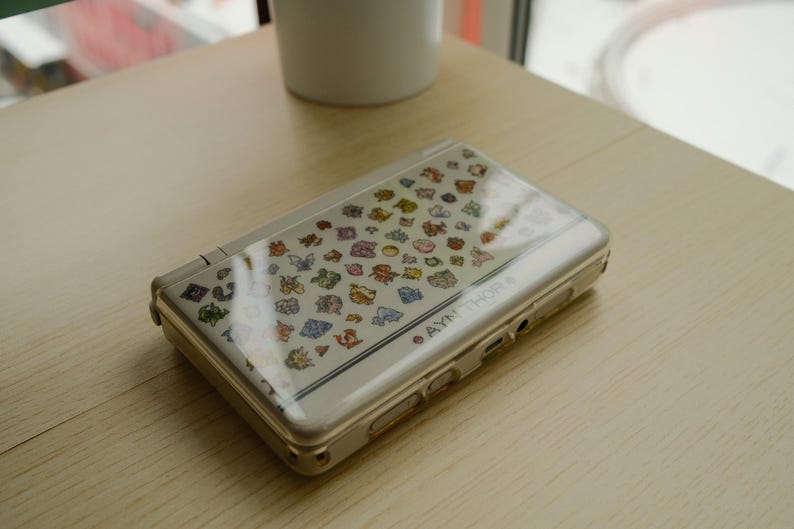 May include: A silver handheld gaming console with a clear cover featuring colorful cartoon character graphics. The console is open and resting on a light-colored wooden surface. The text "AYN THOR" is visible on the side.