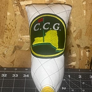 May include: White golf club head cover with a diamond-stitched pattern. The cover features a circular emblem with the letters "C.C.G." and a yellow and green design of a flag and a map.