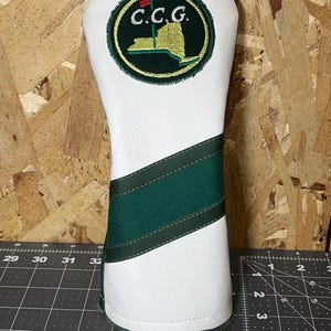 May include: White golf club head cover with a green and white design. The cover features a circular emblem with the letters "C.C.G." and a golf flag over a map of New York State.