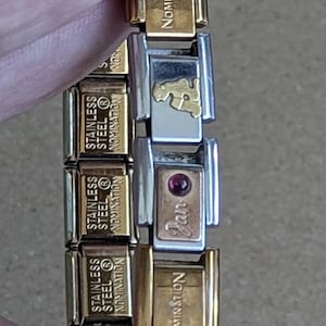 May include: A modular link bracelet featuring gold-tone and silver-tone stainless steel segments. The links are engraved with the word NOMINATION and STAINLESS STEEL. One silver link features a gold-tone dog icon and another has a small red stone.