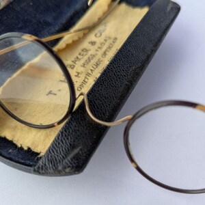 Peut inclure: Lunettes rondes vintage avec monture et branches en écaille de tortue. Les lunettes reposent dans un étui en cuir noir ouvert, avec une doublure en papier jauni. L'étui affiche le nom de l'opticien. Un design classique.