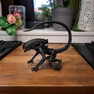 Puede incluir: Una figura negra de Xenomorfo, criatura de la franquicia Alien, posa sobre una superficie de madera. La figura tiene una cola larga y curvada y detalles esqueléticos. La criatura está en posición agachada, con la boca abierta.