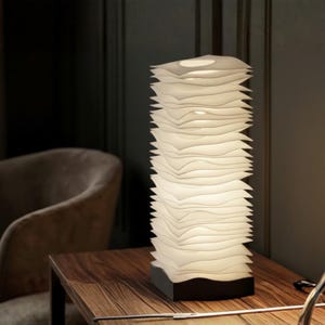 May include: A contemporary white table lamp with a distinctive design. The lamp comprises a stack of layered, wave-shaped components, providing a soft, warm light. It rests on a dark rectangular base, positioned on a wooden table. This lamp is a decorative lighting fixture.