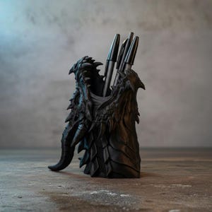 Könnte beinhalten: Schwarzer, drachenförmiger Stifthalter mit Stiften. Der Stifthalter ist aufwendig detailliert und weist Schuppen, Hörner und einen bedrohlichen Ausdruck auf. Die Stifte sind schwarz und elegant und stehen im Kontrast zur strukturierten Oberfläche des Halters. Ein einzigartiges Schreibtischzubehör.