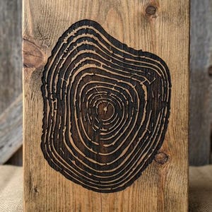May include: A rectangular wooden plaque with a dark, burnt-in tree ring design. The wood has a natural, weathered appearance with visible grain and knots. The tree ring pattern is centered on the plaque, creating a rustic, natural aesthetic.