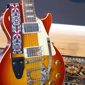 Könnte beinhalten: Eine E-Gitarre mit Sunburst-Finish und einem gemusterten Gurt. Die Gitarre hat ein cremefarbenes Schlagbrett, Chrombeschläge und goldene Knöpfe. Der Gurt ist schwarz mit einem roten und weißen Blumenmuster.