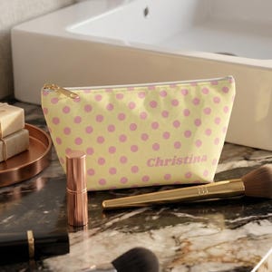 May include: A light yellow zippered pouch featuring a pink polka dot pattern and the name Christina printed in pink. The pouch is accompanied by two makeup brushes, a lipstick, and two stacked bars of soap on a copper-colored tray.