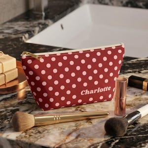 May include: A maroon cosmetic pouch featuring a white polka dot pattern and the name Charlotte printed in white script. The pouch has a gold-toned zipper closure and is positioned alongside two makeup brushes, a lipstick, and two stacked soap bars.