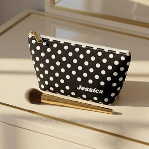 May include: A black cosmetic pouch featuring a white polka dot pattern and the name Jessica printed in white on the front. The pouch has a white zipper with a gold-toned metal pull. A makeup brush with a gold handle lies in front of the pouch.