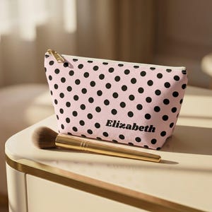 May include: A light pink cosmetic pouch featuring a black polka dot pattern and the name Elizabeth printed in black lettering on the front. The pouch has a gold-toned zipper closure and is accompanied by a makeup brush with a gold handle.